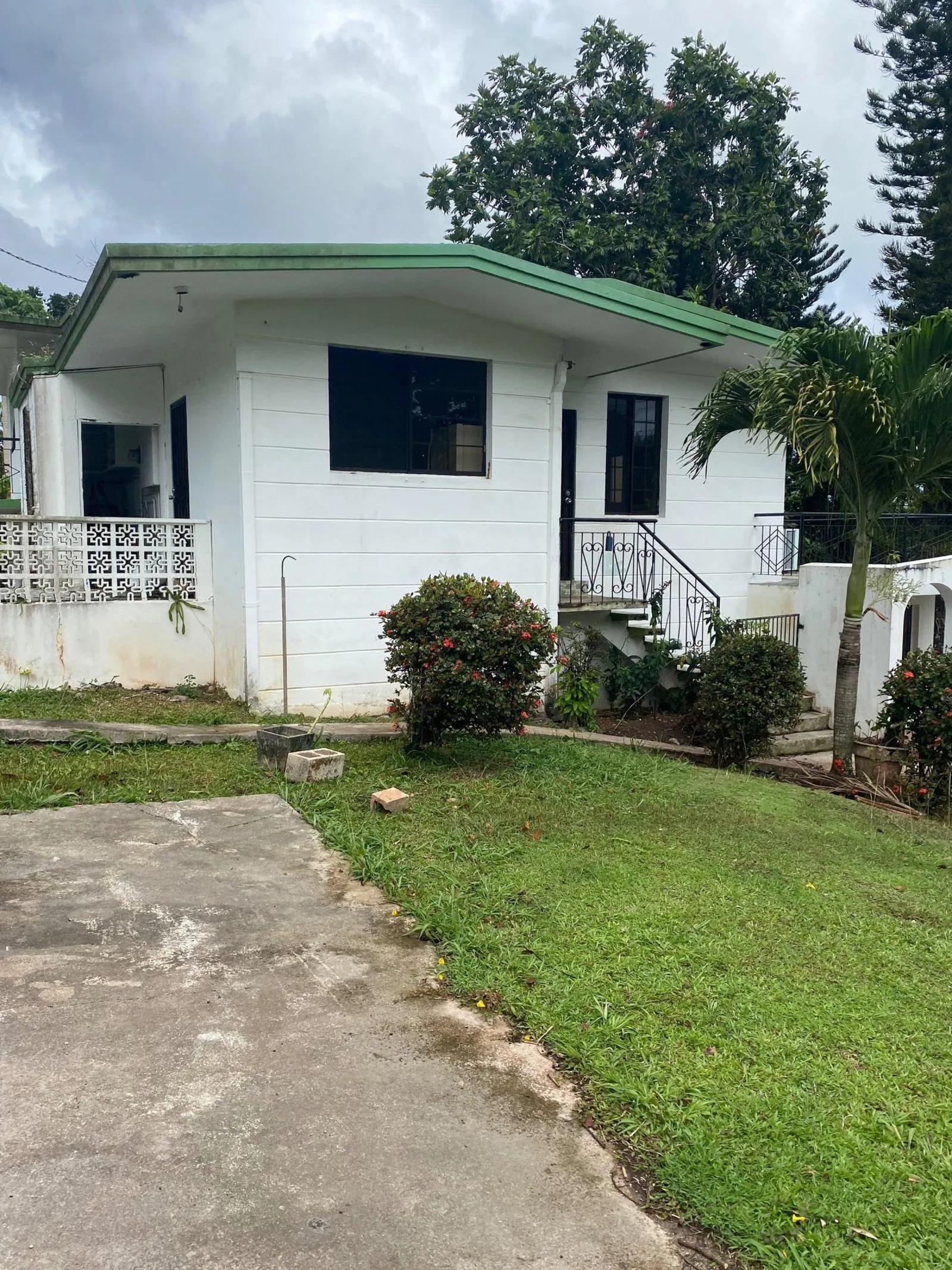 An older single-storey Guam concrete house with a green roof edge, breeze-block porch screen and weather-stained walls